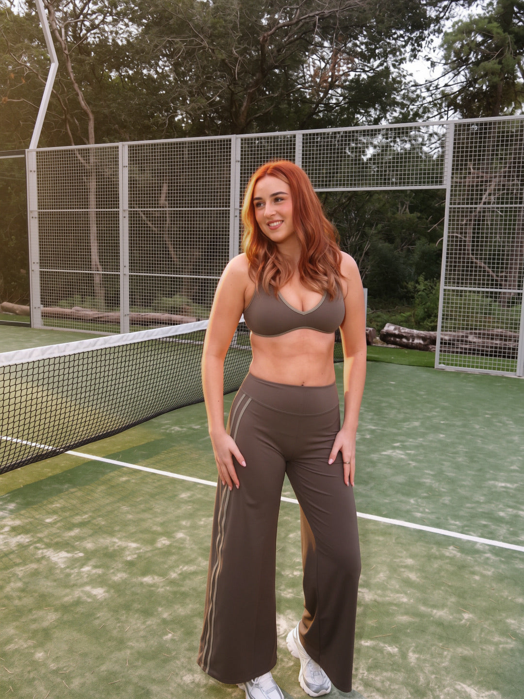 A woman with red hair stands on an outdoor tennis court wearing a matching brown activewear set consisting of a sports bra and wide-leg pants.