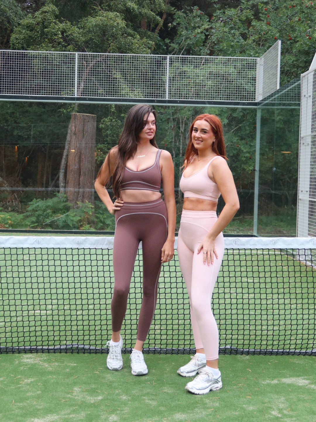 Two women posing in matching athletic wear on an outdoor tennis court. The woman on the left wears a dark brown sports bra and leggings set, while the woman on the right wears a light pink set.
